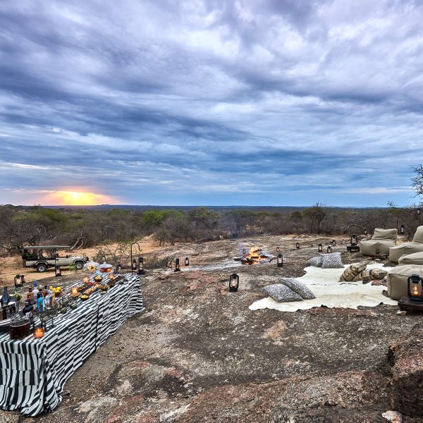 Blankets, poofy chairs, lanterns, and a long table with drinks and snacks set up for sundowners on the big boulders by Mwiba Lodge