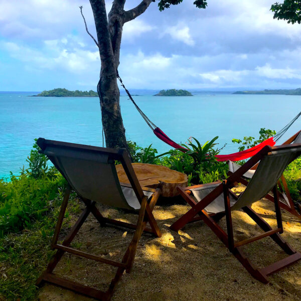Red hammock hanging from two trees with two deck chairs overlooking the ocean and islands in the distance